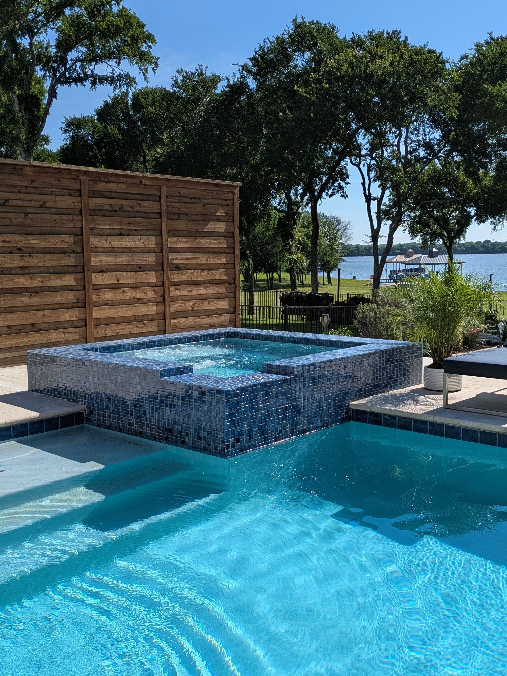 Outdoor pool and hot tub with blue tile, overlooking a lake and trees.