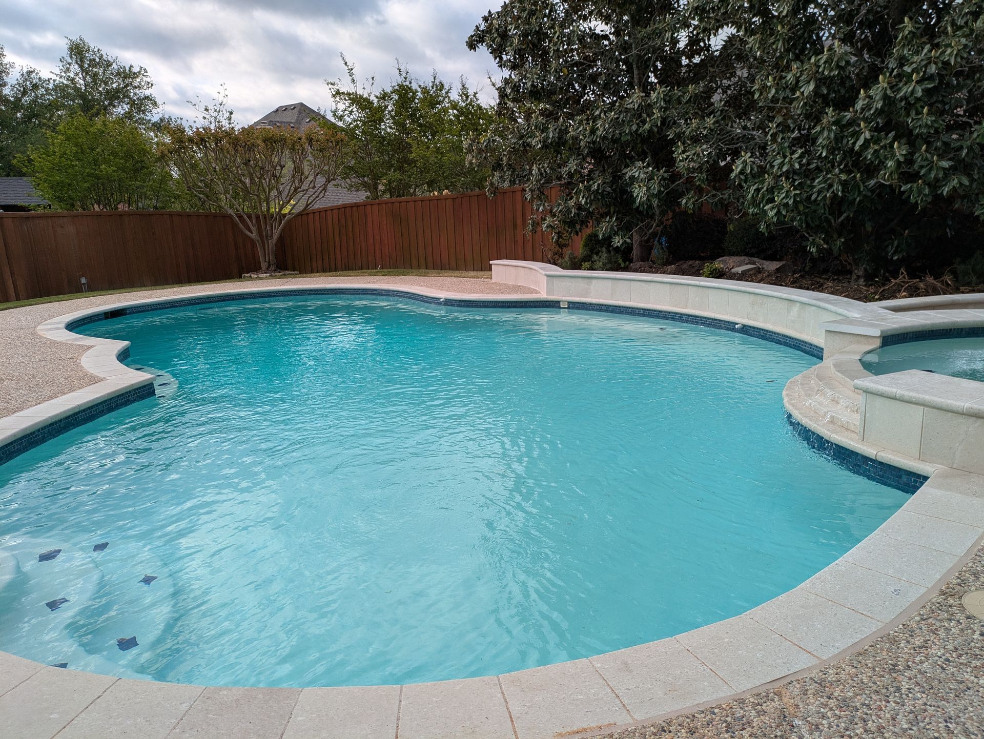 Swimming pool with turquoise water, stone coping, and a spa. Brown wooden fence and trees in background.