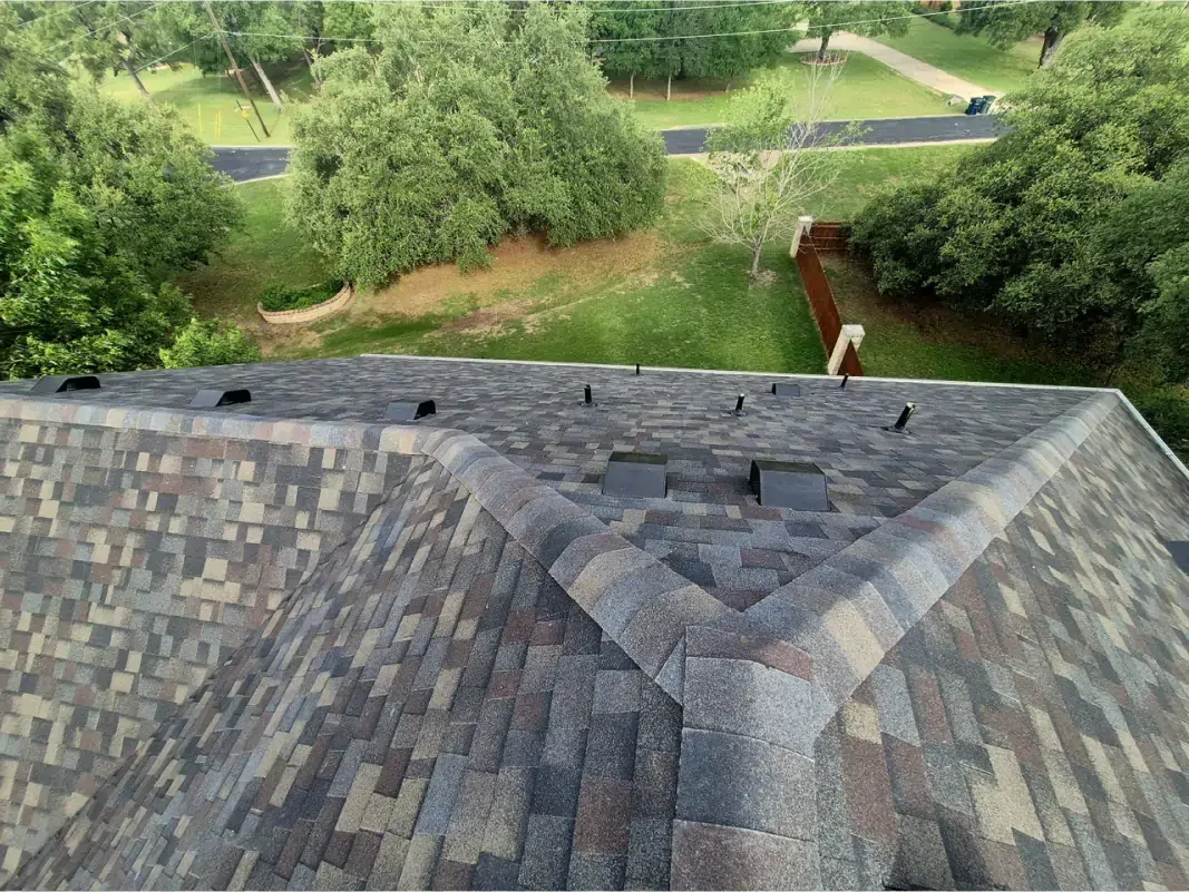 Overhead view of a roof with brown, gray, and blue shingles surrounded by green trees and grass.