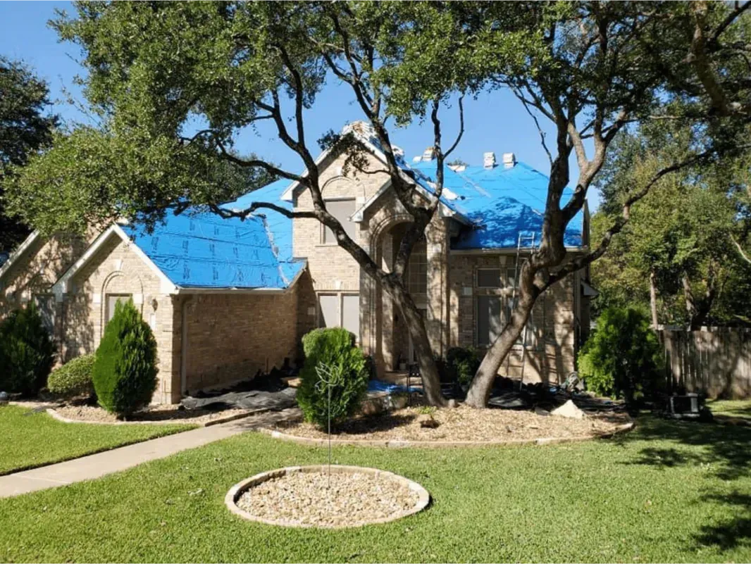 House with blue tarp covering a damaged roof, surrounded by green lawn and trees.
