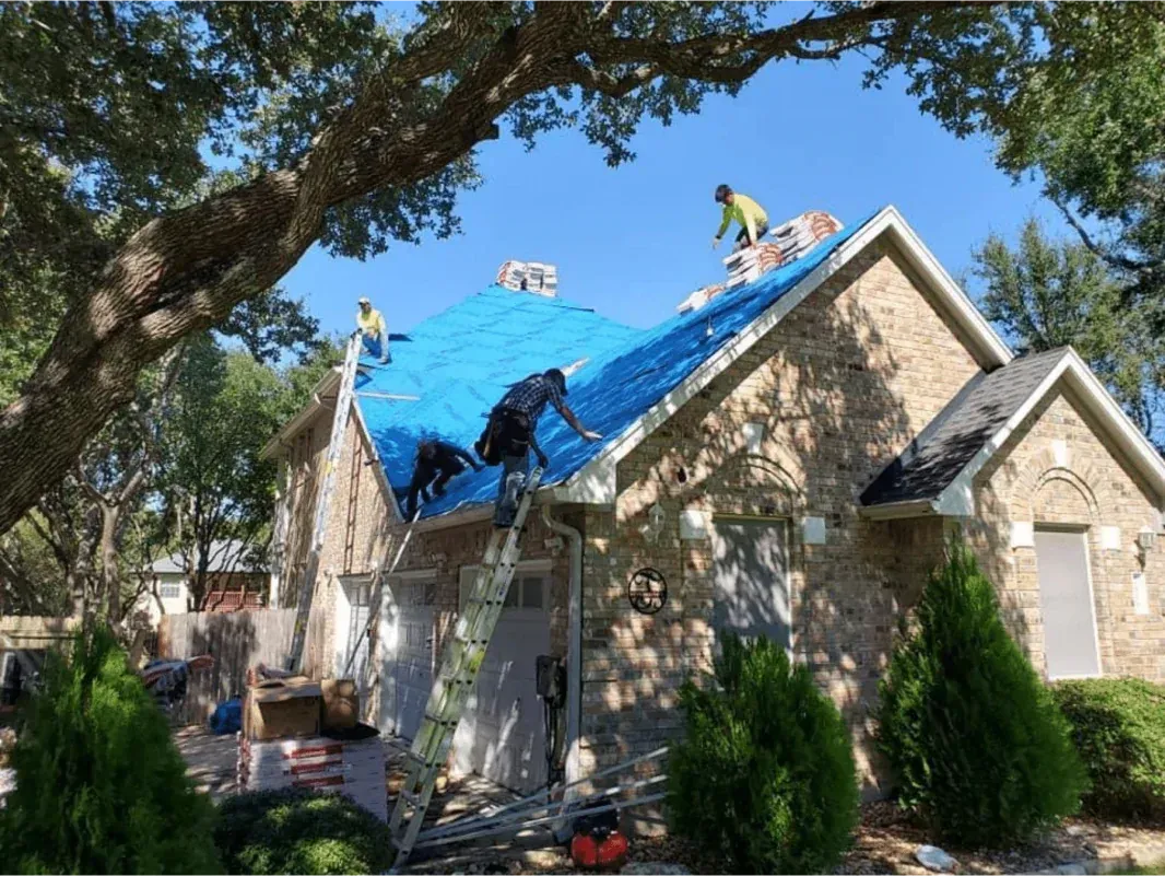 Roofers repairing a two-story house with a blue tarp, surrounded by trees, on a sunny day.