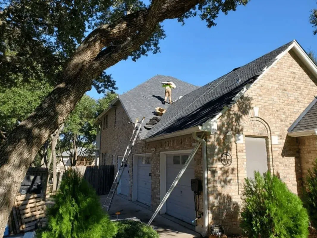Roofer on a gray shingled roof of a two-story brick house with a ladder, working under a tree on a sunny day.