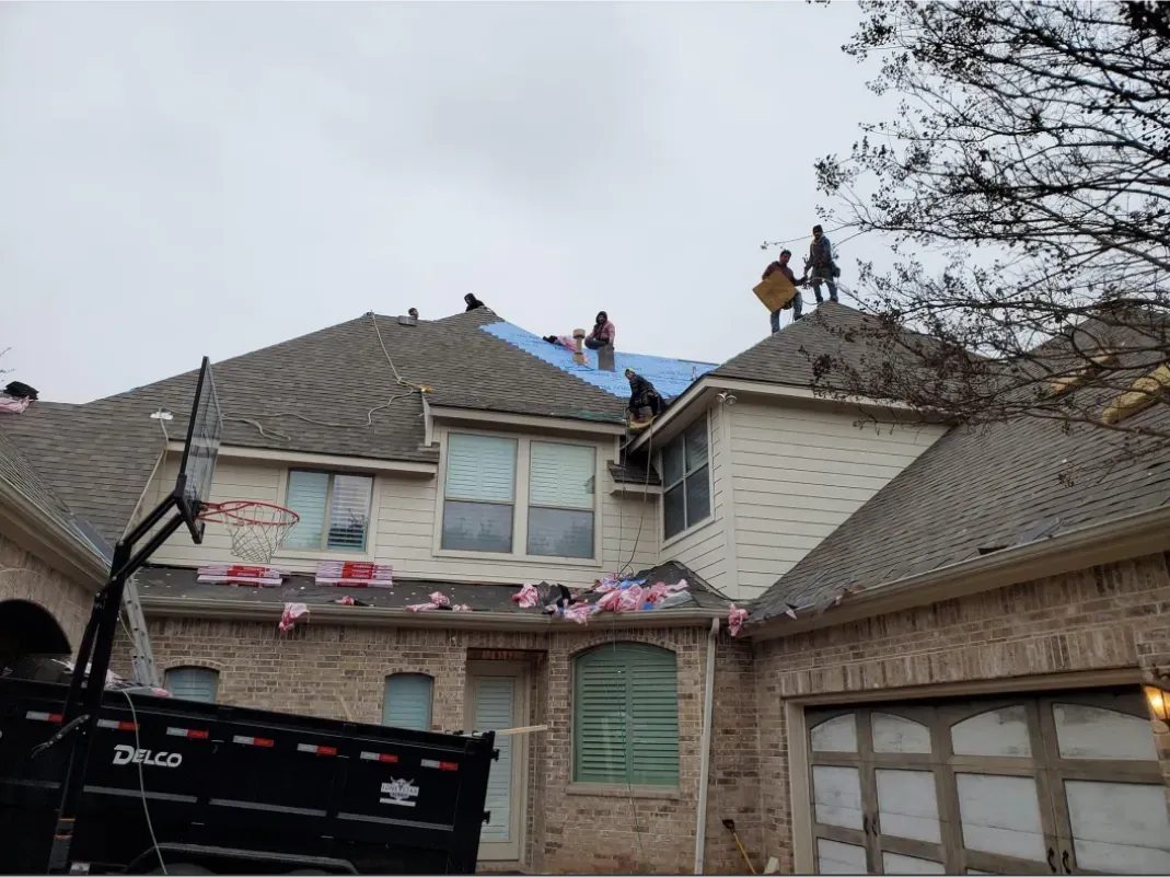 Roofers working on a house under a cloudy sky. Blue tarp covers a section. Black trailer in front.