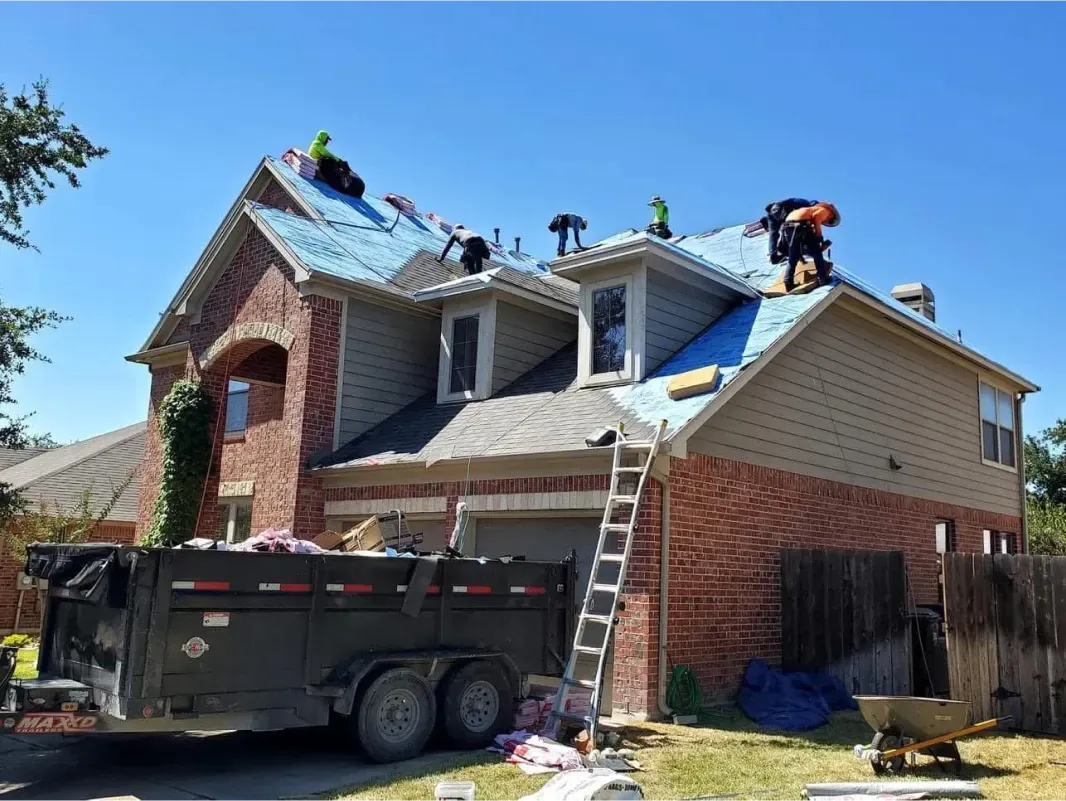 Roofers installing blue underlayment on a two-story house under a bright, sunny sky, with a dumpster in front.