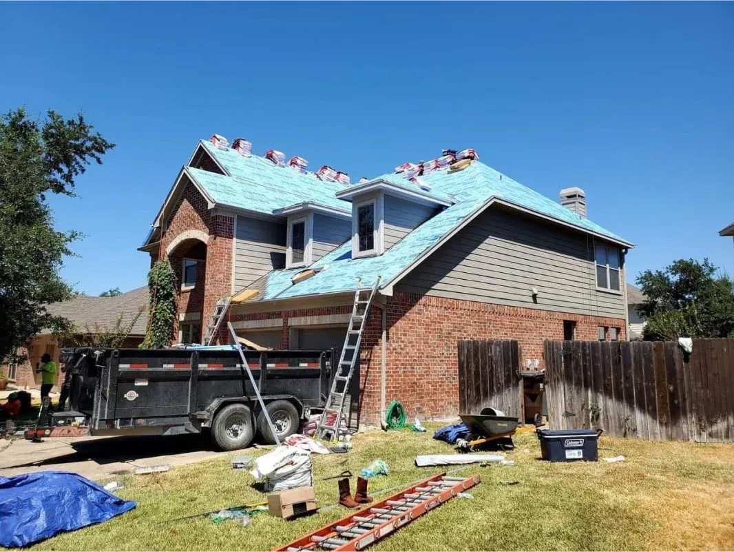 Residential home under construction with new roofing and materials, blue tarp, and trailer in yard.