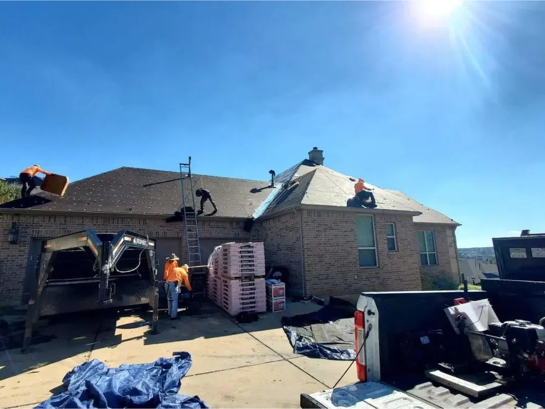 Roofers working on a brick house roof under a bright blue sky. Pink insulation and a ladder are visible.