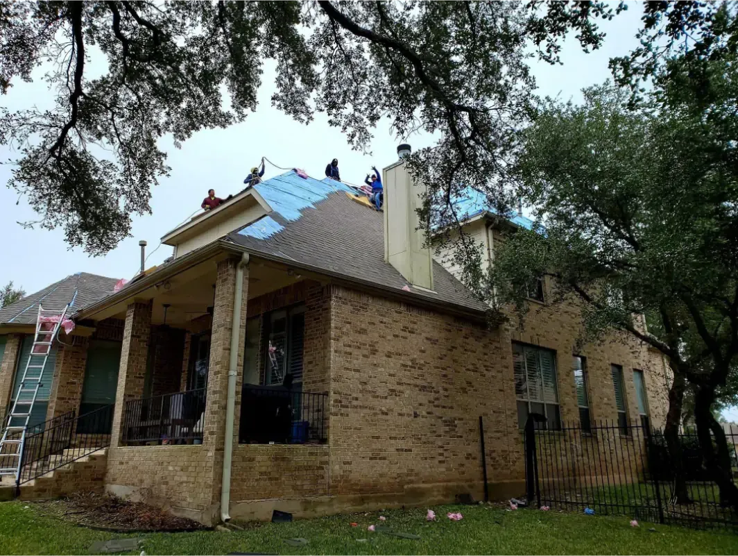 Roofers working on a house with brown brick exterior and blue tarp. Overcast sky.