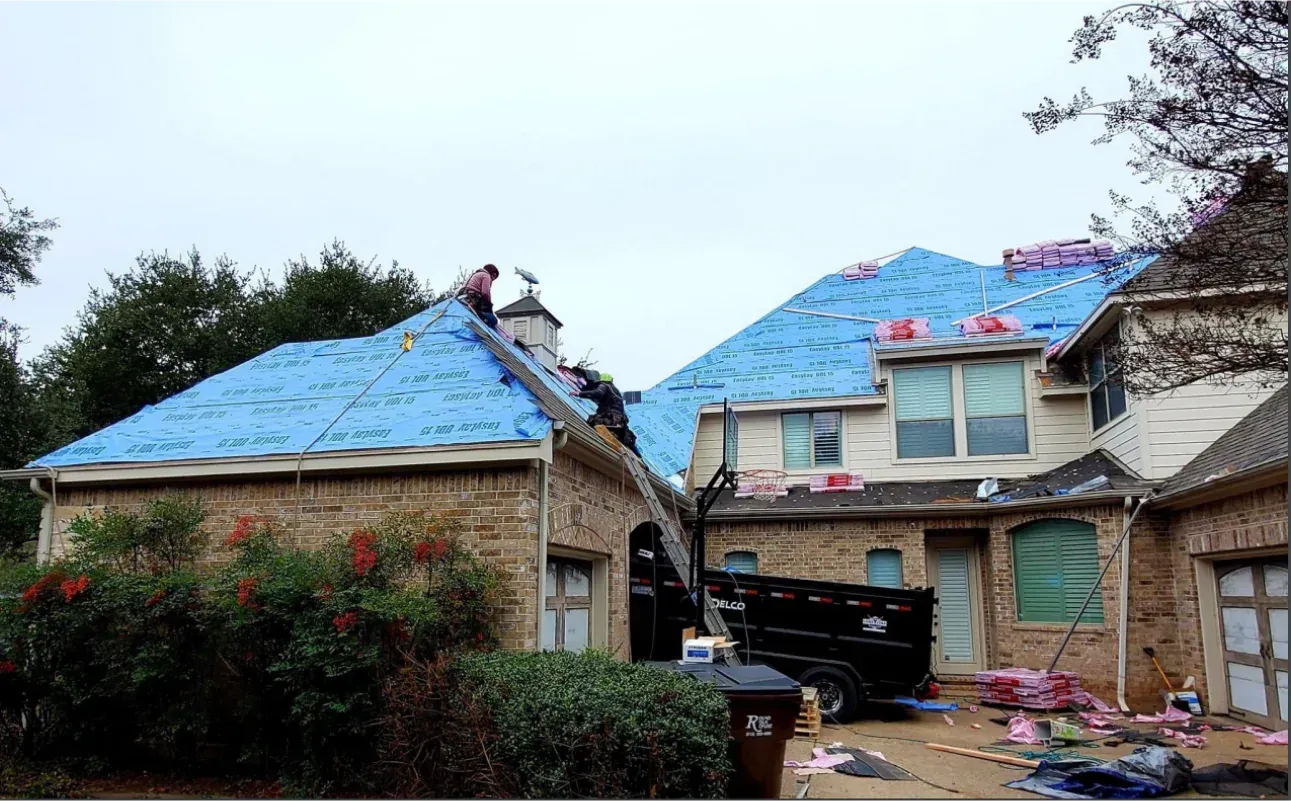 Roofing work on a two-story brick house; blue tarp, workers on roof, cloudy sky.