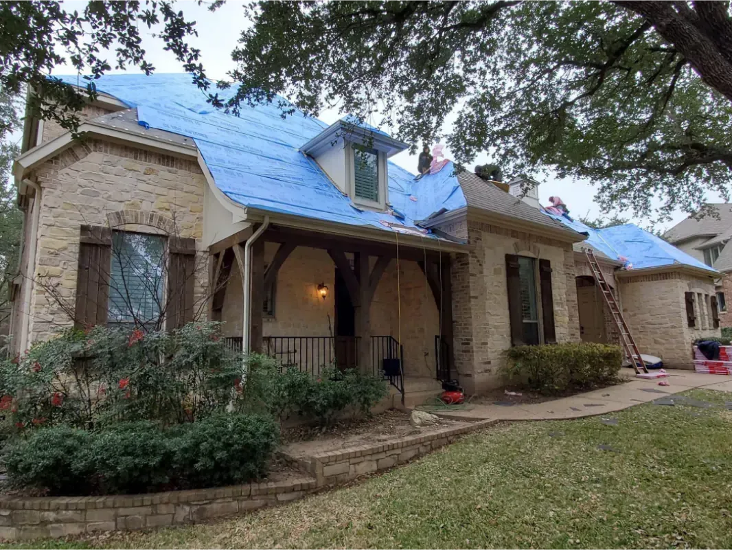 House with blue tarp on roof, two workers, brown brick, cloudy day.