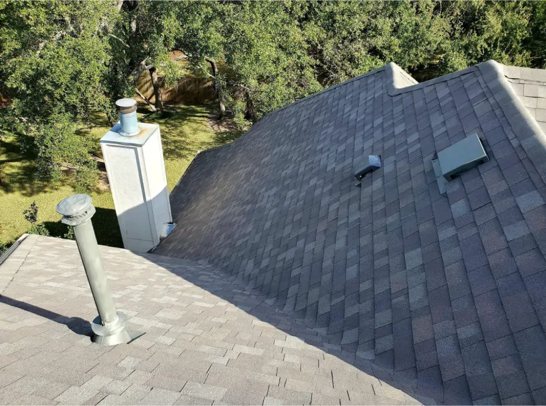 Gray shingled roof with a chimney and vents against a backdrop of green trees.
