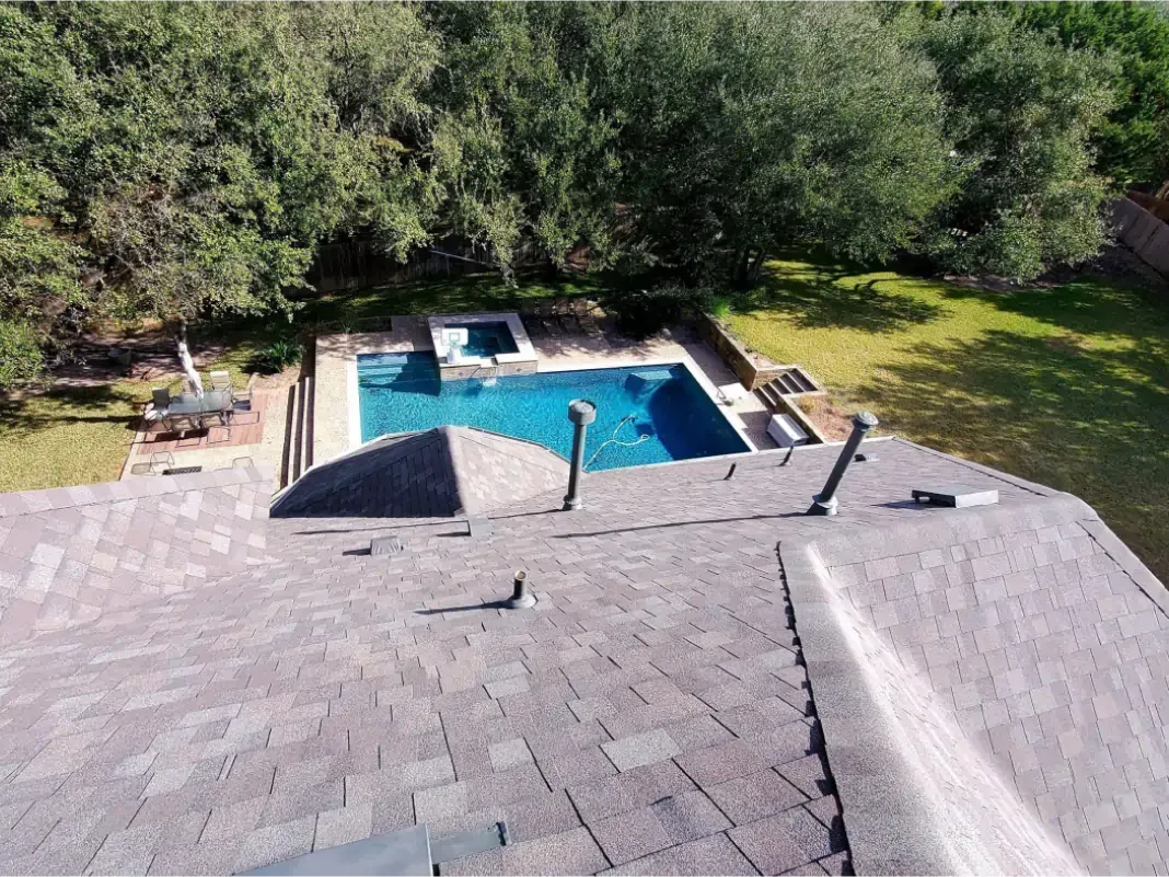 Overhead view of a house roof and backyard pool with surrounding trees and grass.