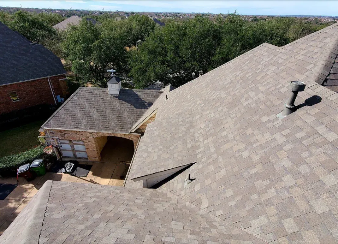 Overhead view of a house with brown shingled roof and brick siding, trees and sky in the background.