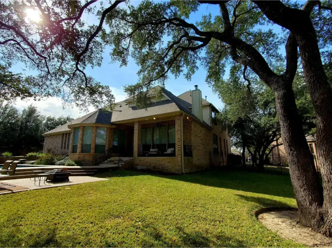 House exterior with a covered patio, green lawn, and large tree in the foreground under a sunny sky.