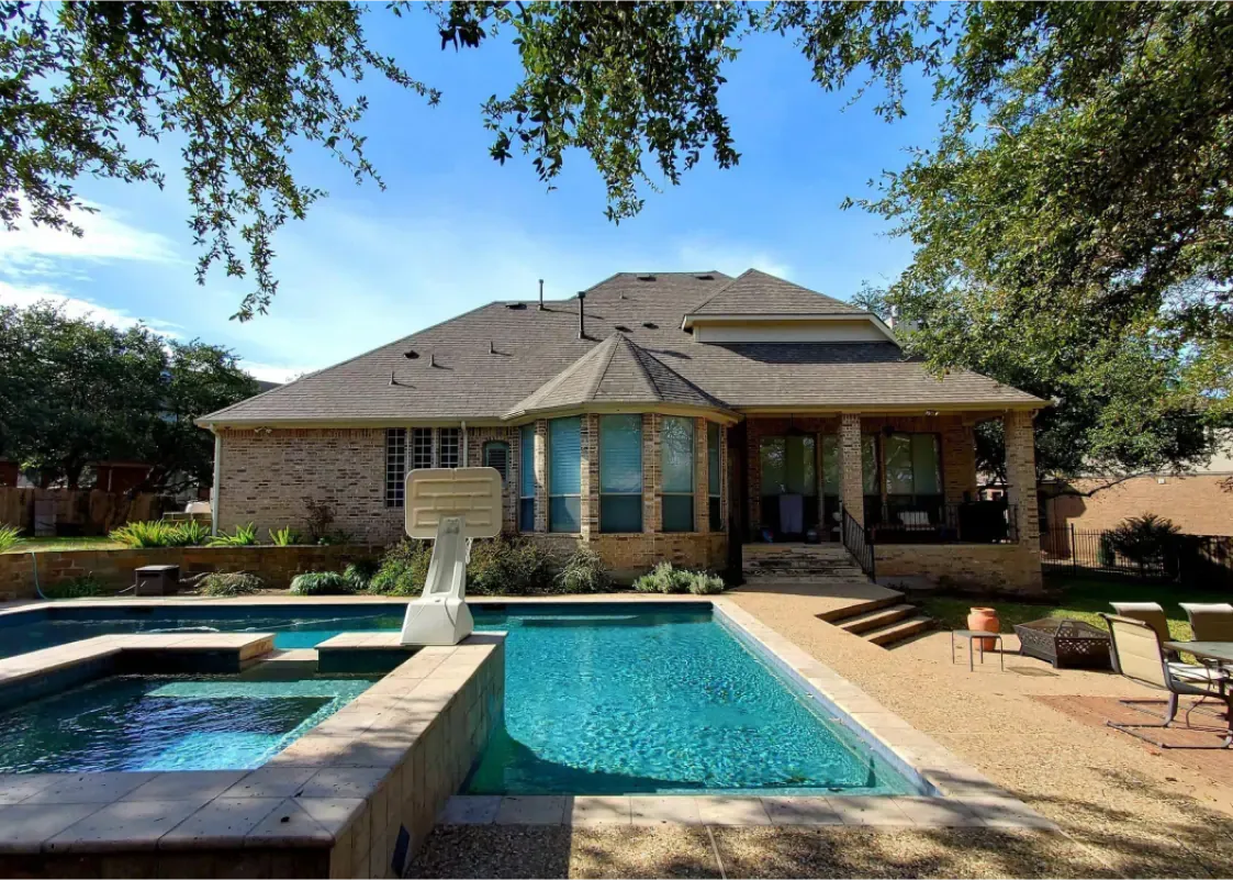 Backyard with a pool and spa. Brick home with a covered patio. Blue sky with trees overhead.