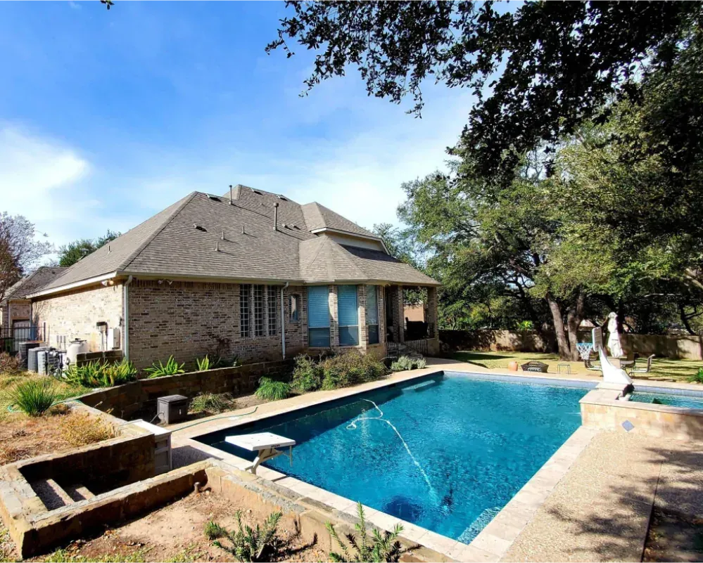 Backyard with a swimming pool next to a brick house under a bright blue sky.