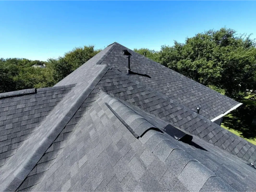 Dark shingled roof with vents and ridge cap, viewed from above against a clear blue sky and trees.
