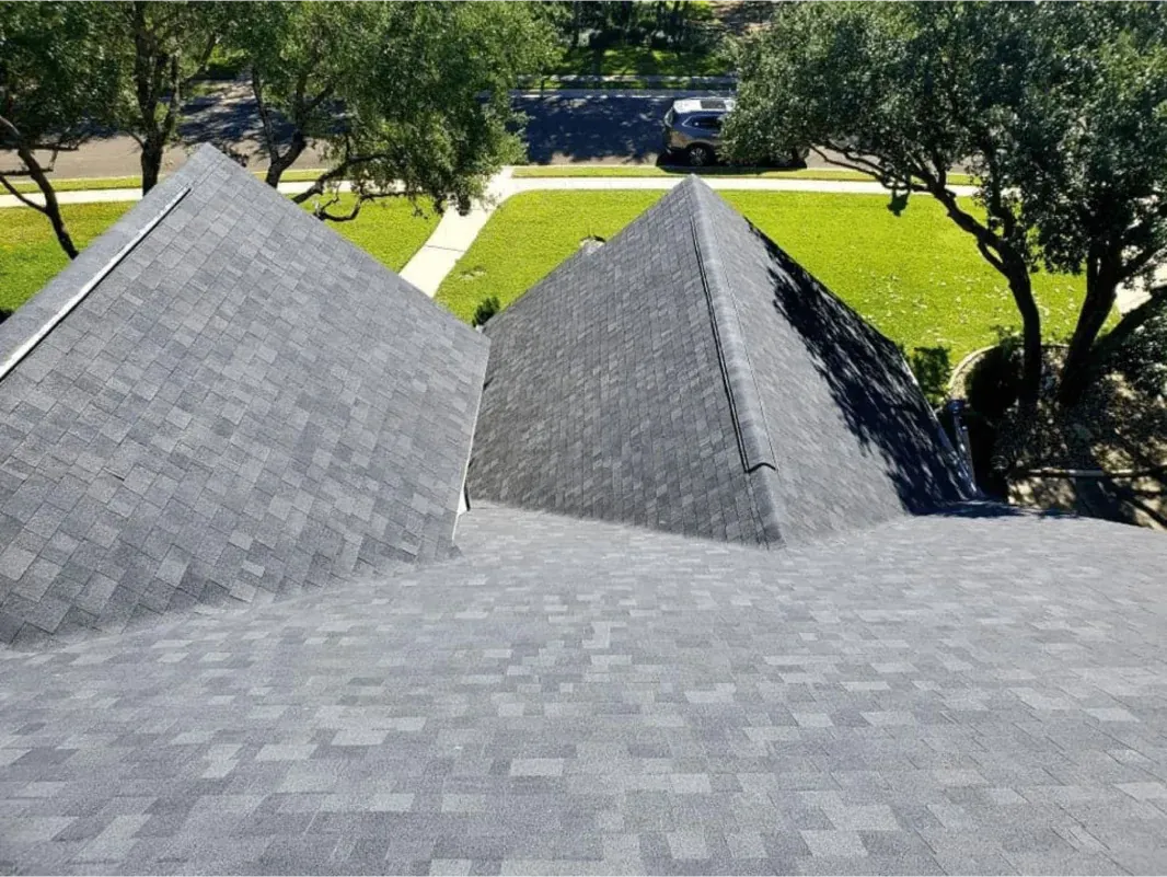 Gray shingled roof peaks with a grass yard and trees in the background.