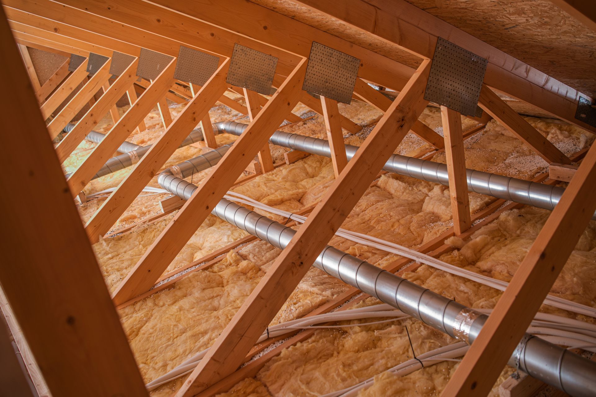 Attic interior featuring wooden roof trusses, exposed metal ventilation ducts, and yellow fiberglass insulation.