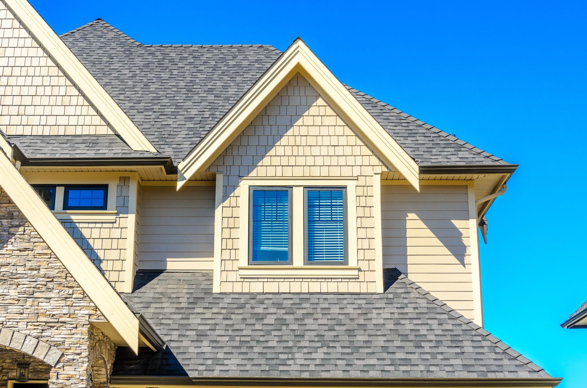 Close-up of a house with a gray shingled roof, beige siding, and a blue sky. Close-up of a house with a gray shingle roof and a beige dormer with windows against a blue sky.