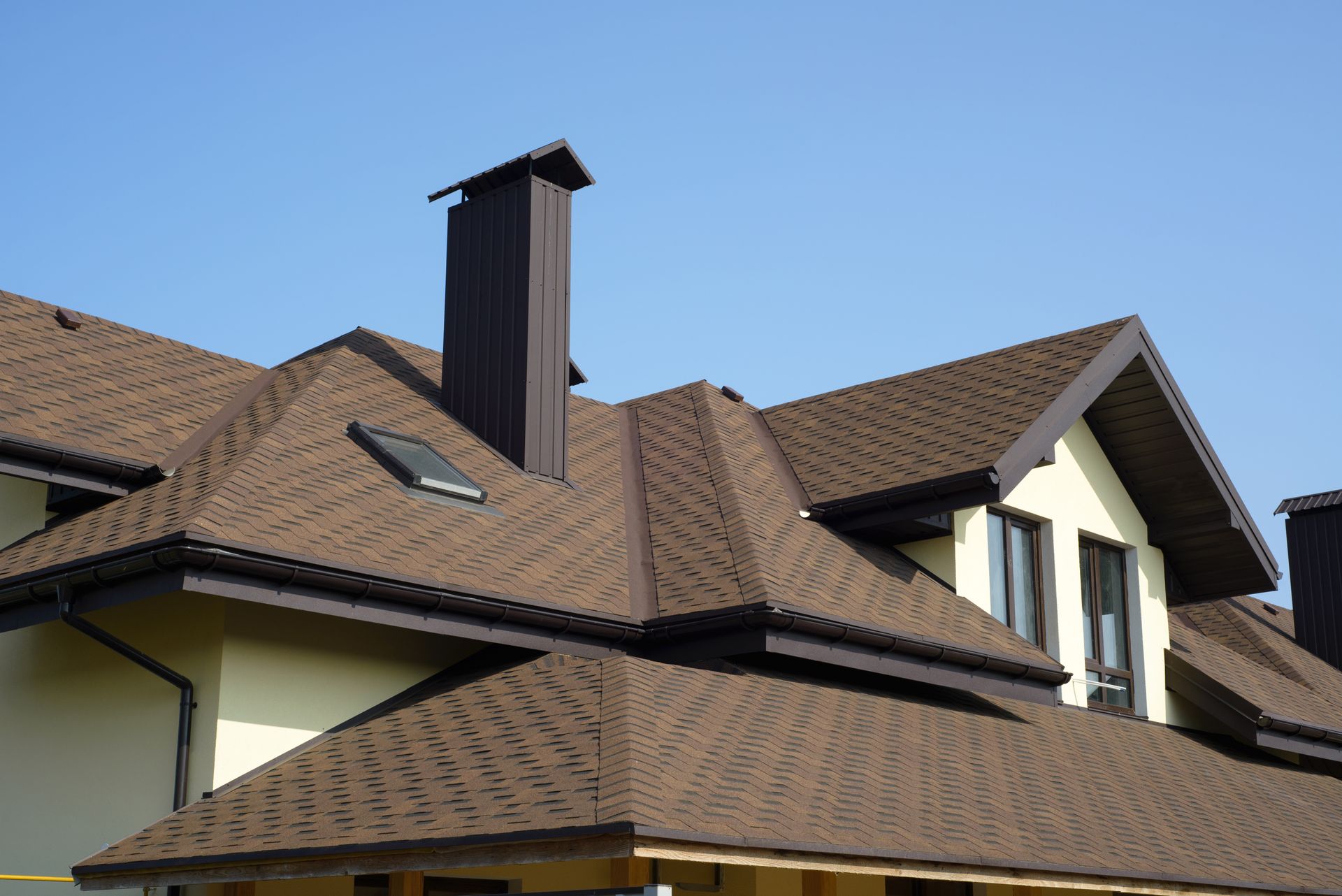 Brown shingled roof with a chimney and dormer windows against a clear blue sky.