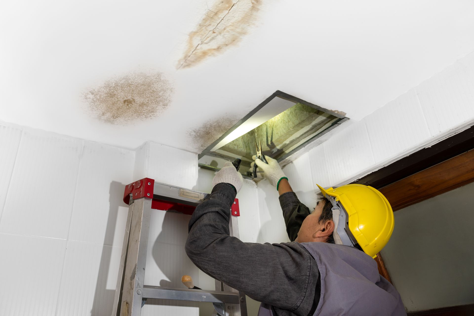 Person in yellow hard hat inspecting ceiling ventilation, water damage present.