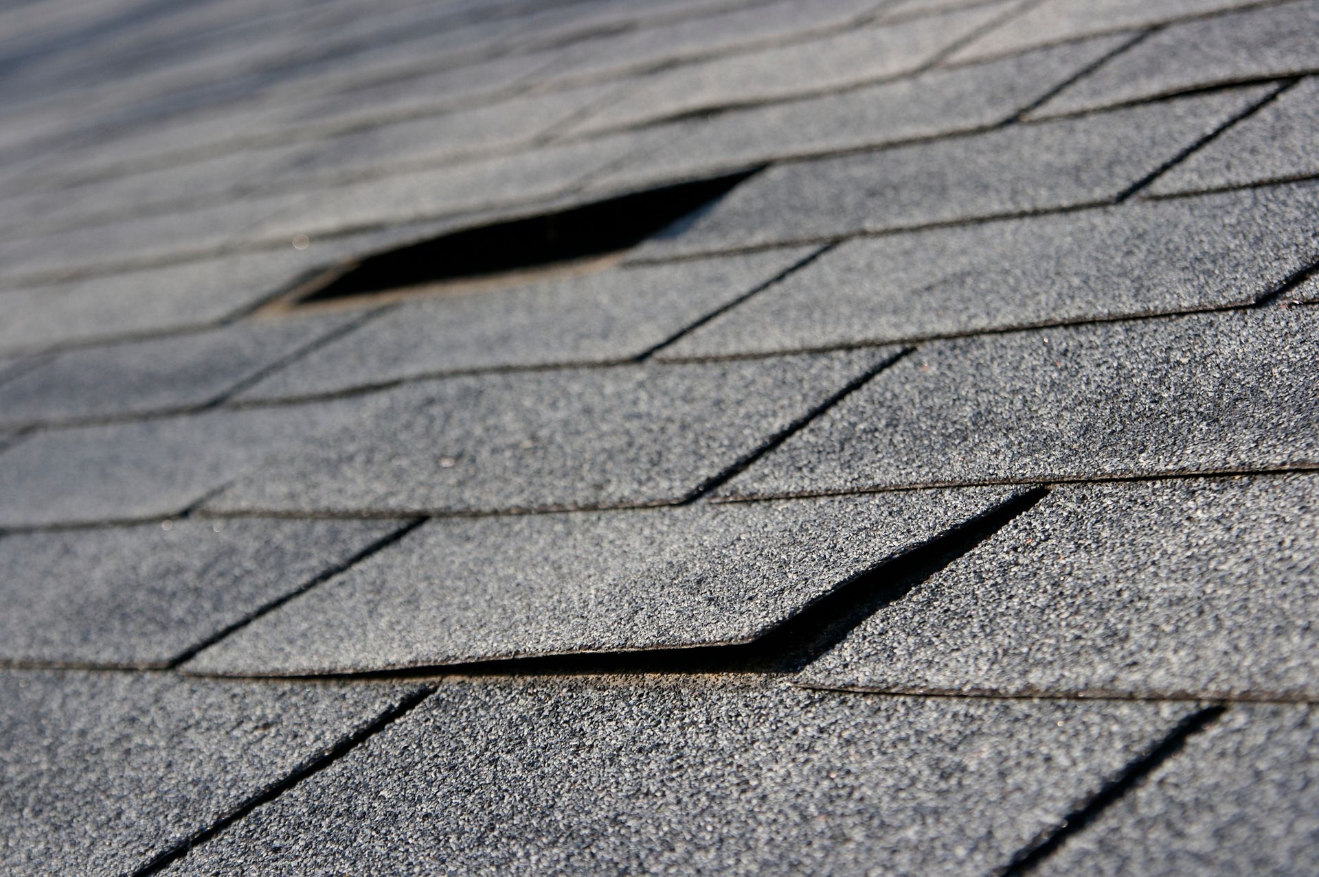 Close-up of a gray asphalt shingle roof with a hole and lifted shingles.