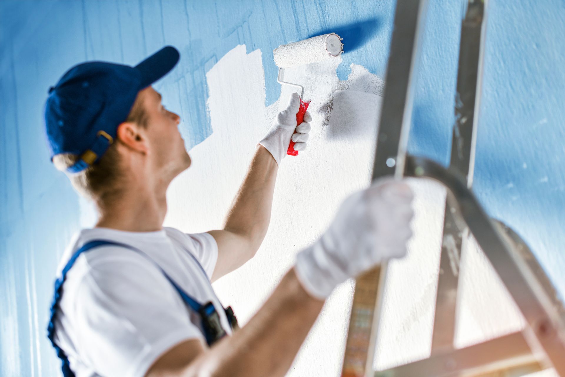 Man in overalls and cap painting a blue wall white with a roller while standing on a ladder.