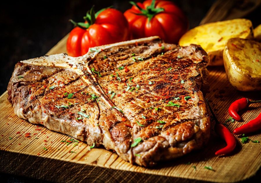 A large steak is sitting on a wooden cutting board next to tomatoes and potatoes.