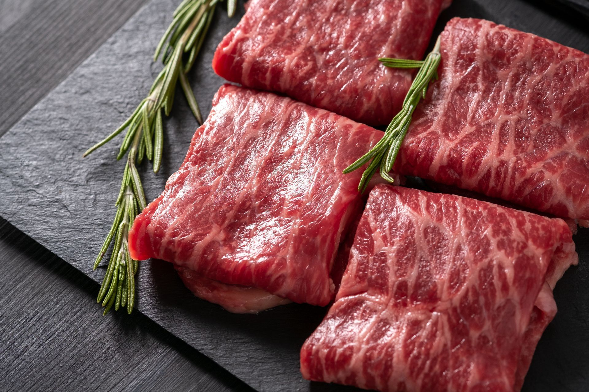 Four pieces of raw beef with rosemary on a cutting board.