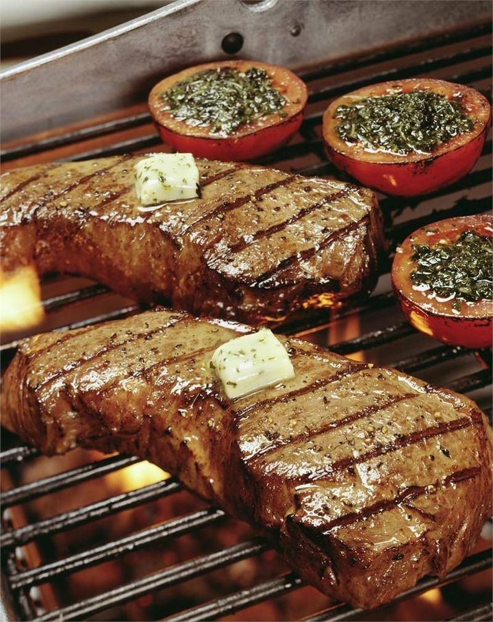 Steaks and tomatoes are cooking on a grill with butter on top