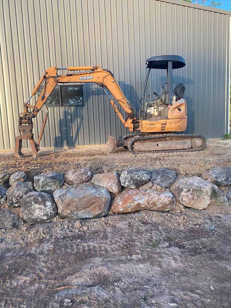 A Small Excavator is Parked in Front of a Building Next to a Pile of Rocks — Blue Gum Earthworks in Atherton, QLD
