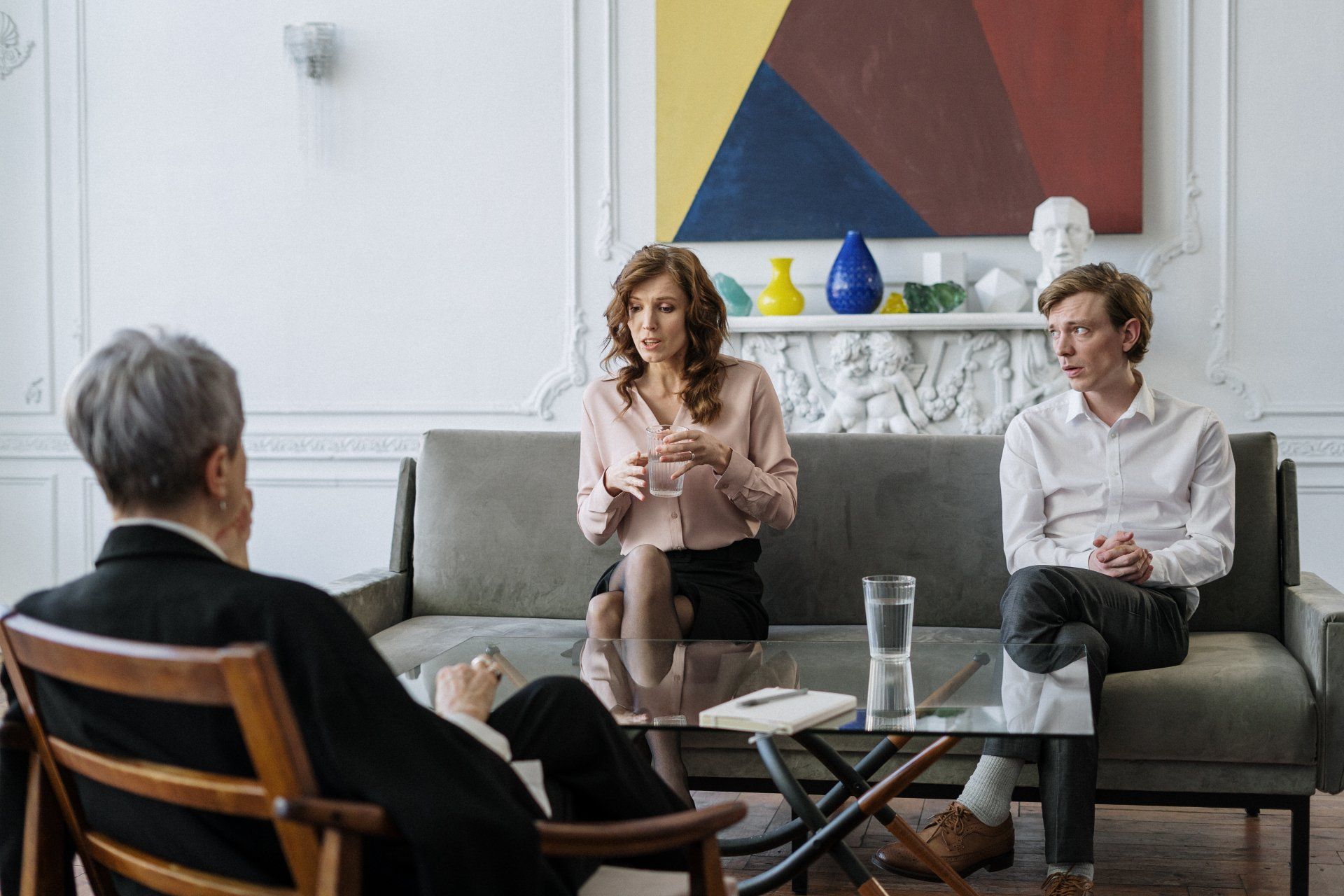 Three people in a living room setting during a discussion, with one seated facing two others on a sofa.