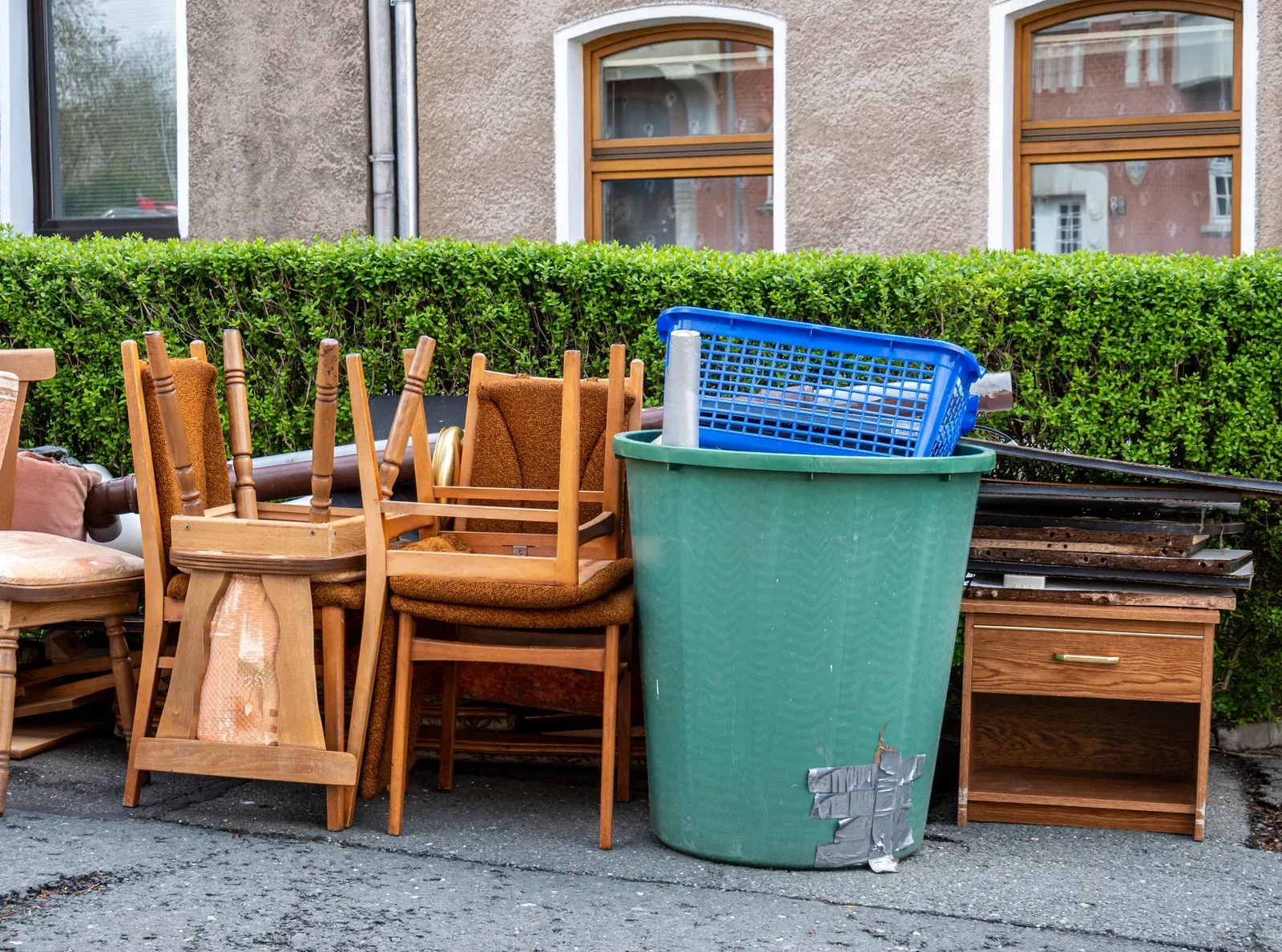 A pile of wooden furniture and a green trash can on the side of the road.