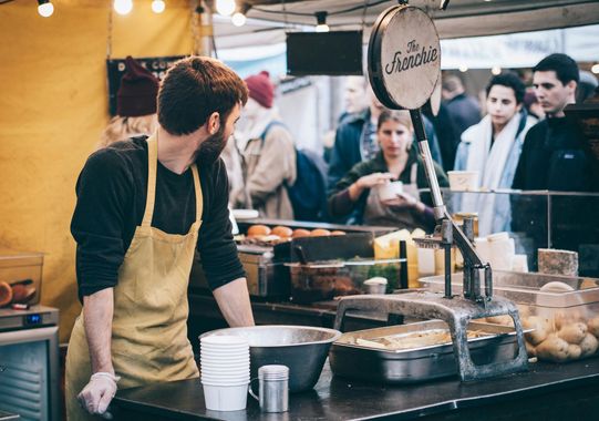 Man in yellow apron at a food stall, serving customers. People in background, food and containers on the counter.