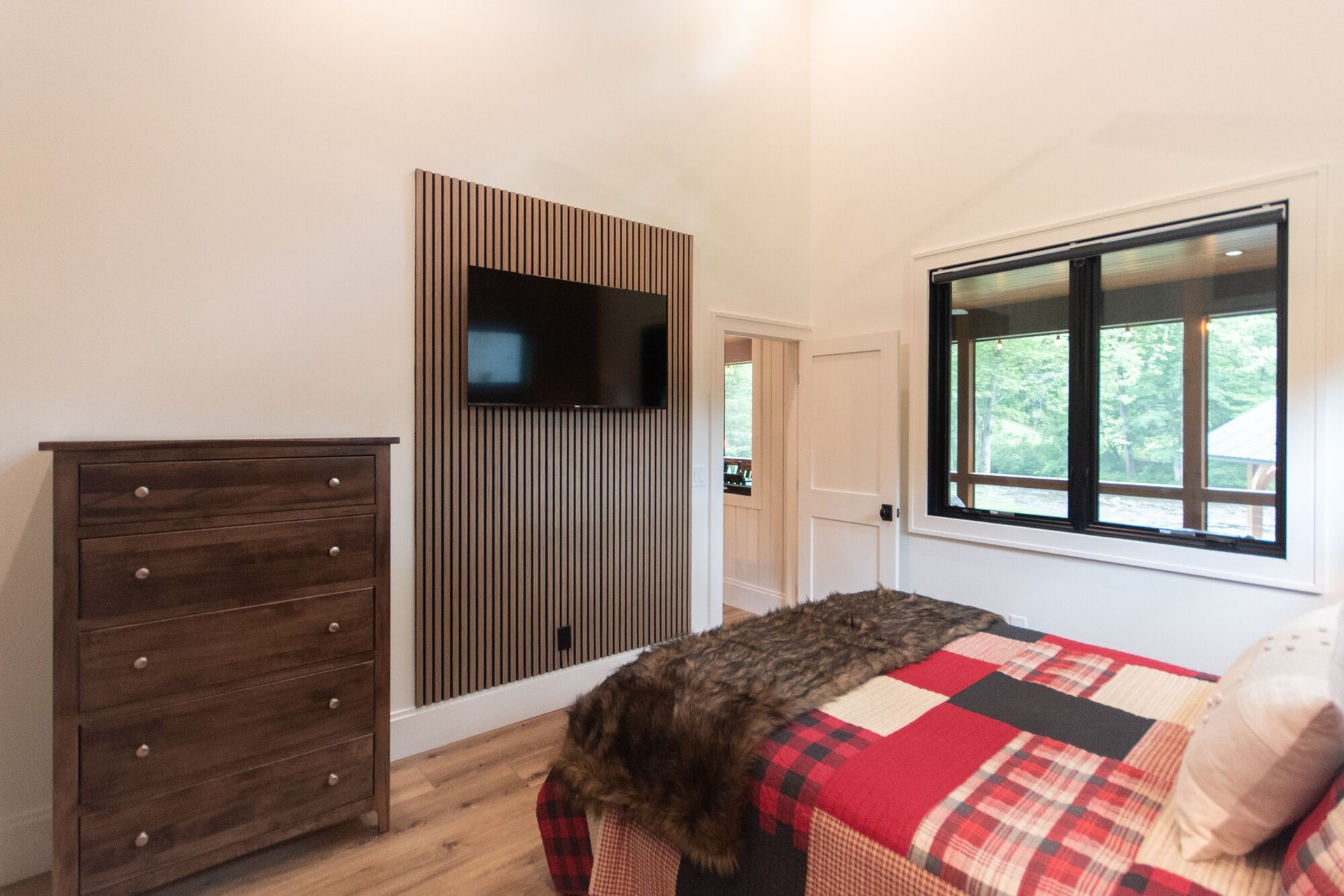 Bright bedroom with a bed, dresser, wall-mounted TV, wood slat accent, and large window.