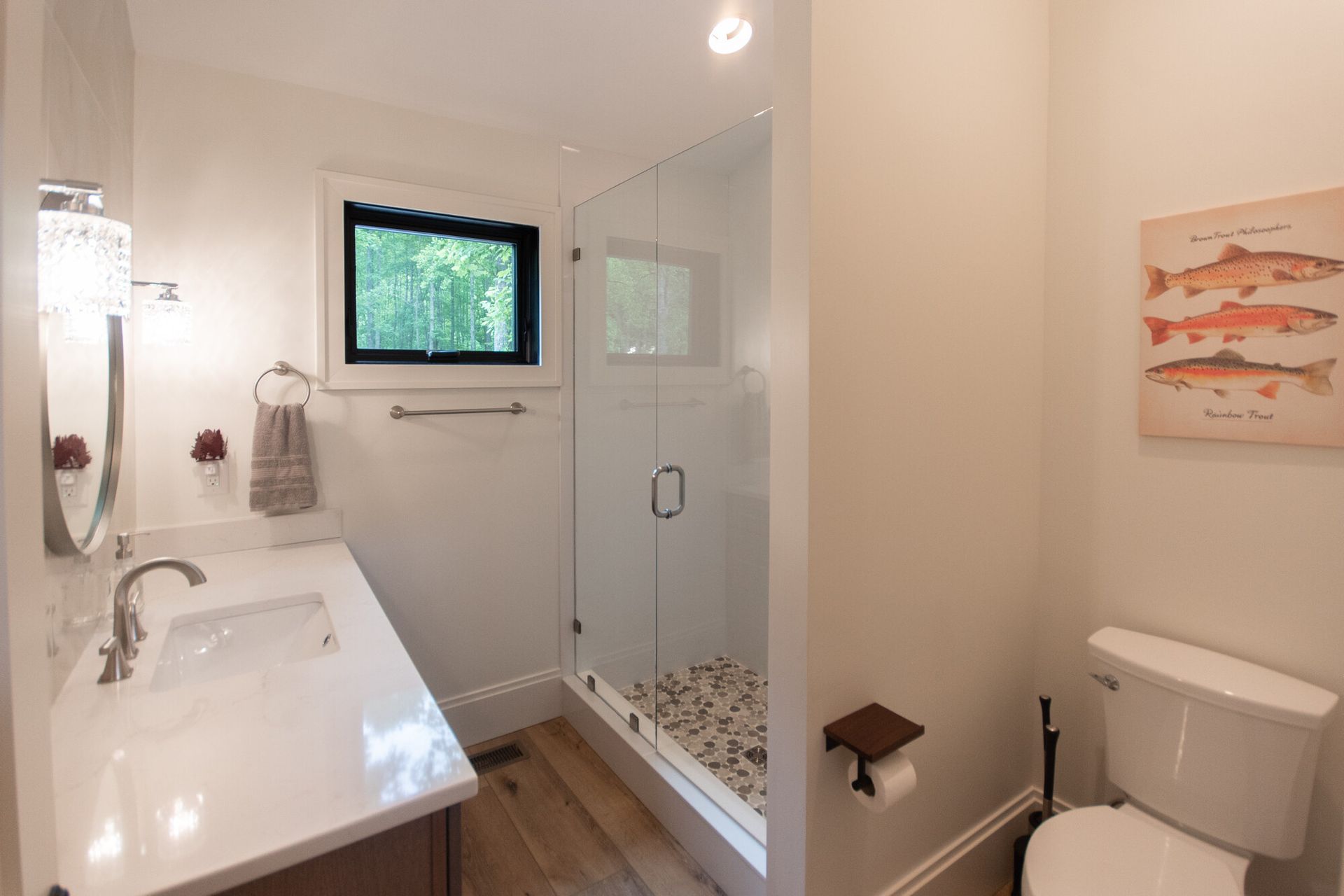 Bright bathroom with white sink, glass shower, toilet, and small window over wood floor.