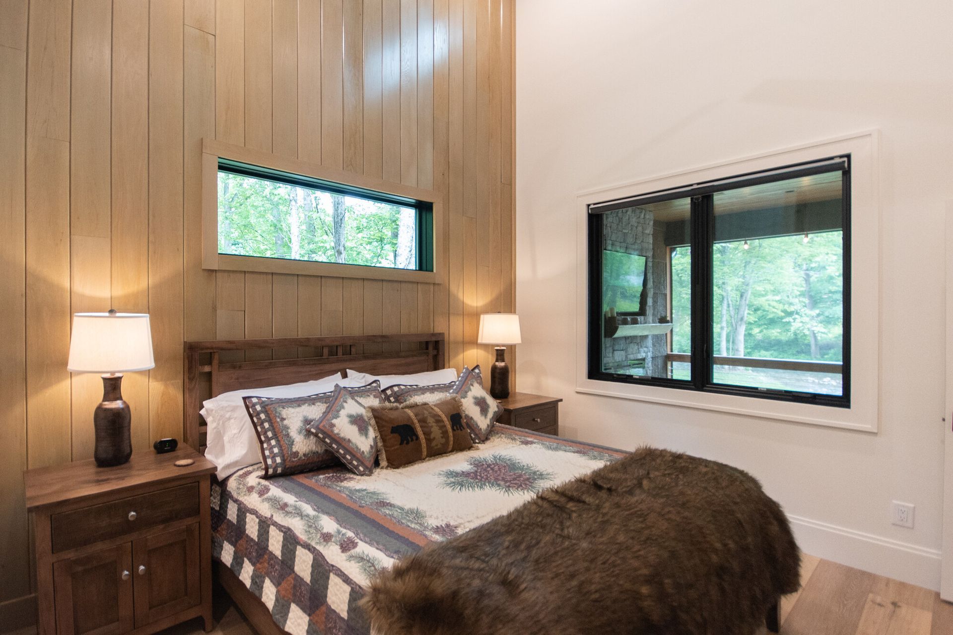 Cozy bedroom with wood accent wall, bed, bedside lamps, and large windows overlooking trees