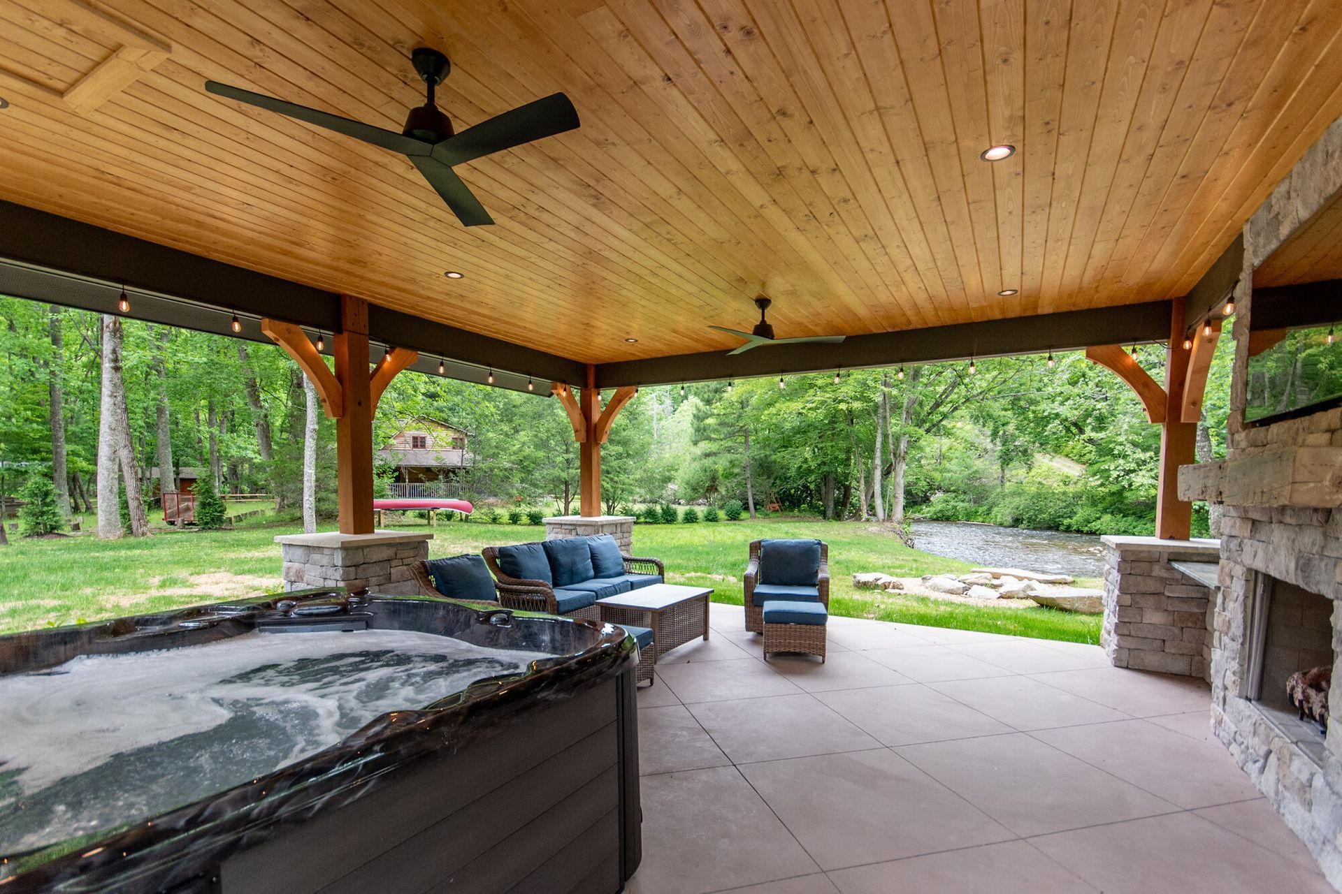 Covered outdoor patio with wood ceiling, ceiling fan, stone fireplace, seating, and green backyard view