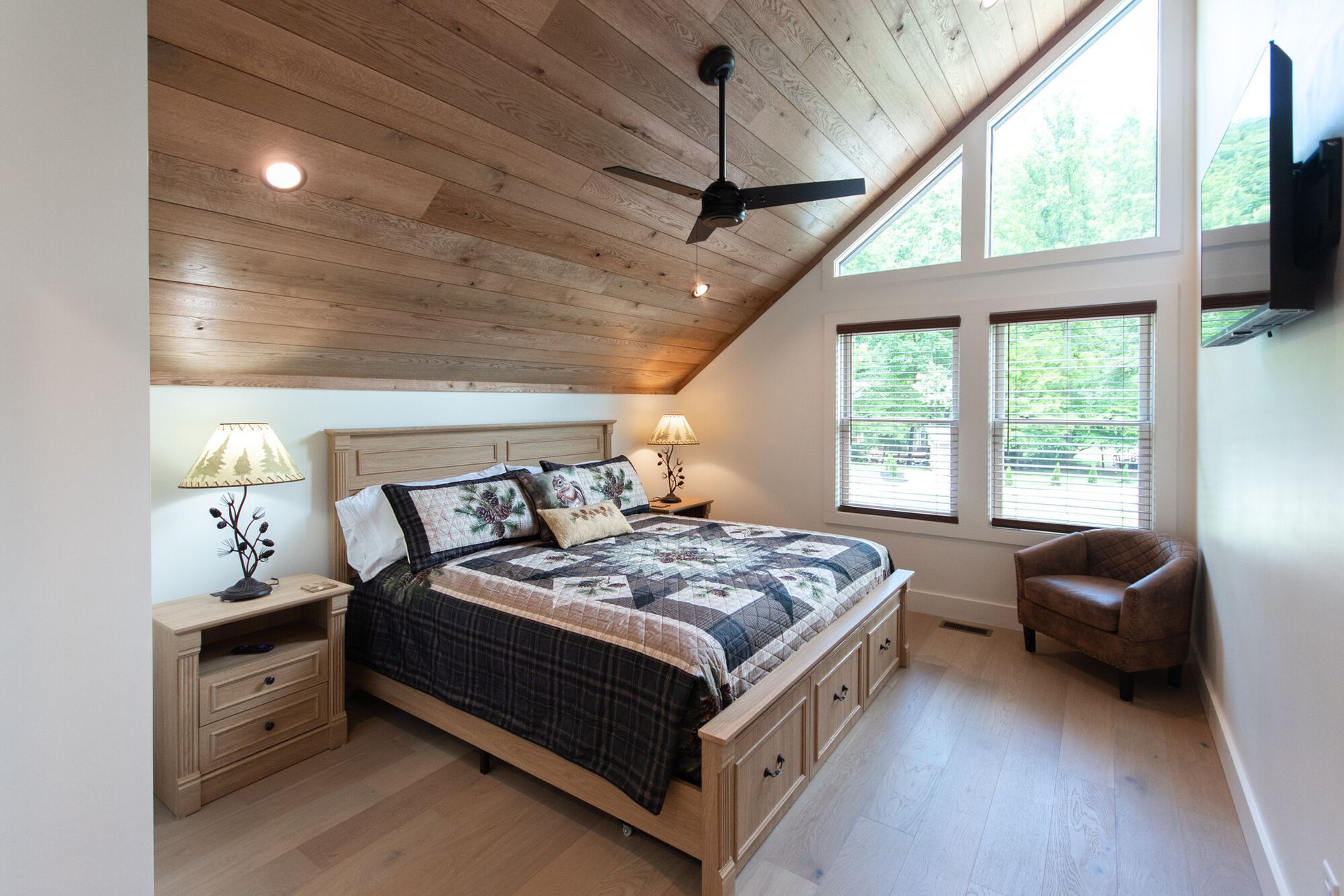 Cozy attic bedroom with wood-paneled ceiling, large windows, and a bed with patterned bedding