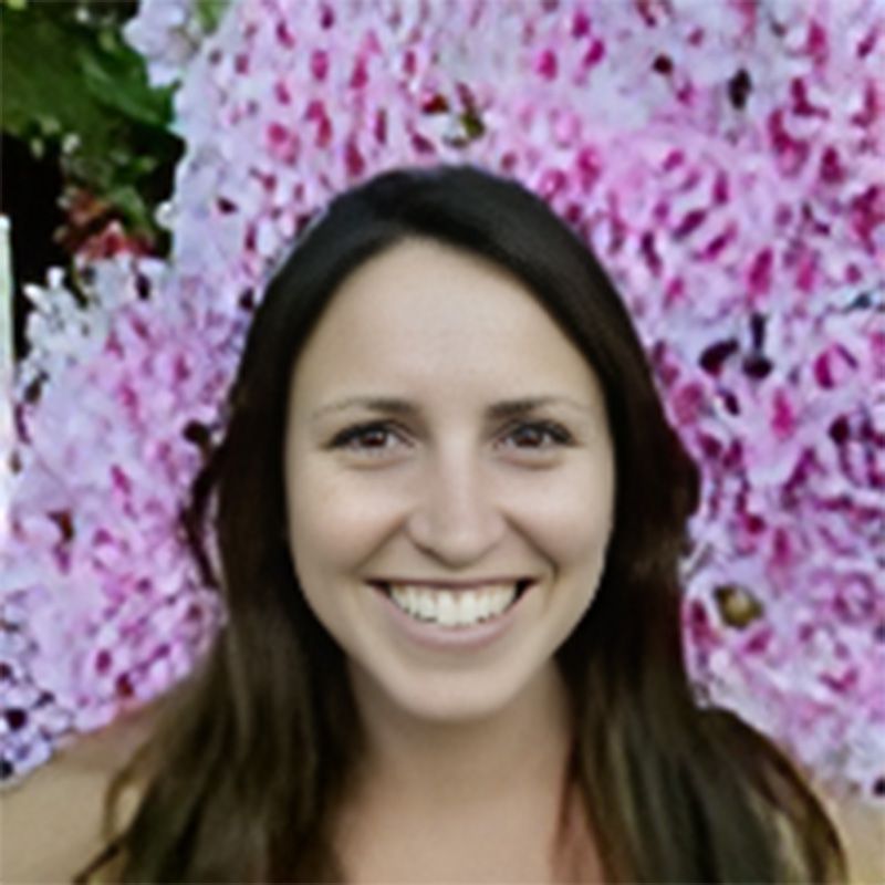A woman is smiling in front of a wall of pink flowers.