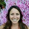 A woman is smiling in front of a wall of pink flowers.