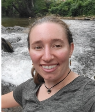 A woman is smiling in front of a wall of pink flowers.