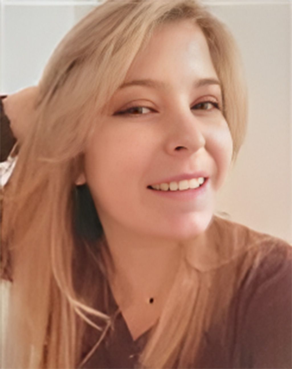A woman with long blonde hair is smiling and holding her hair.