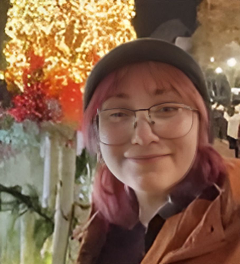 A woman with pink hair and glasses is smiling in front of a christmas tree.