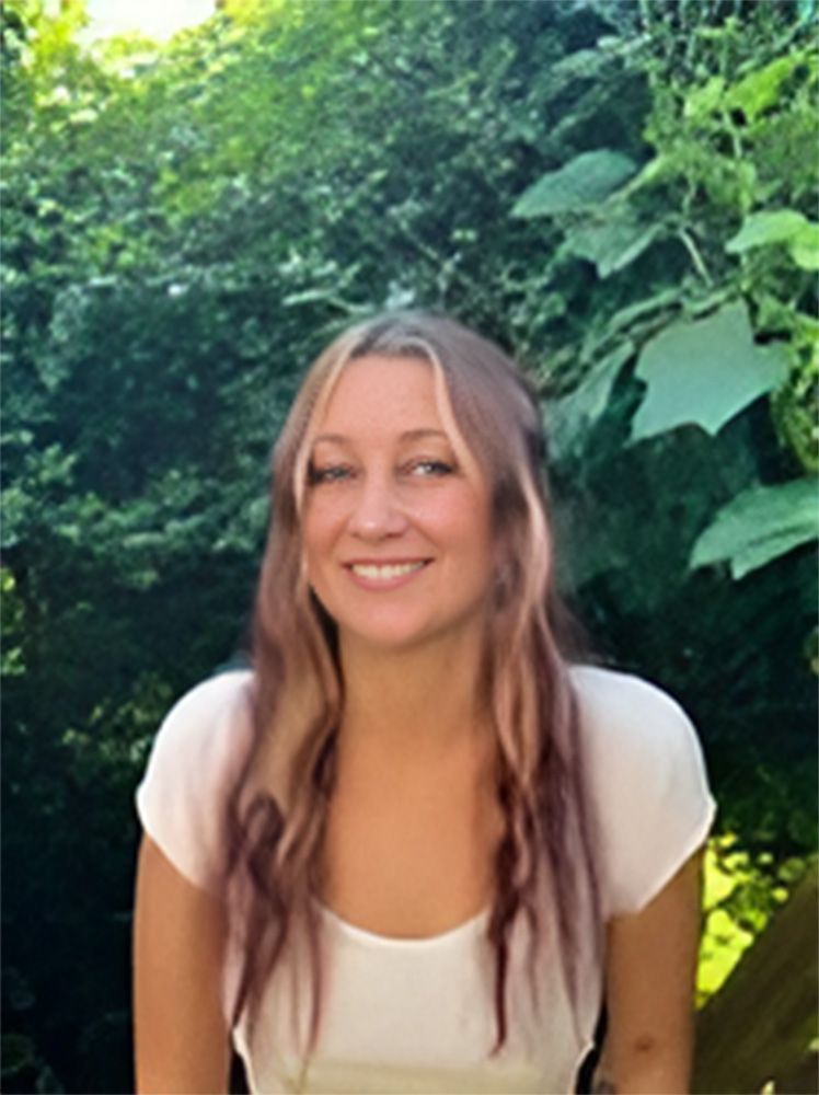 A woman in a white shirt is smiling in front of a tree.