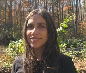 A woman is smiling in front of a wall of pink flowers.