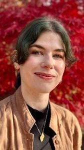 Woman with dark hair smiles, wearing a tan jacket, black shirt, and pendant; red foliage background.