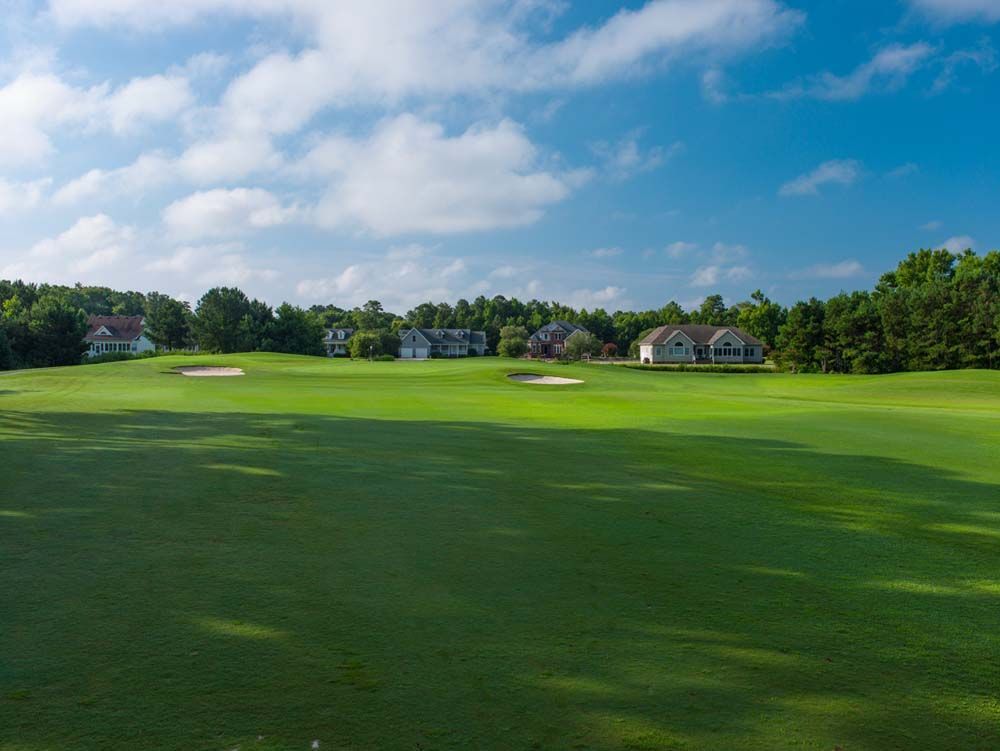 Green golf course with houses in the background under a blue sky with white clouds.