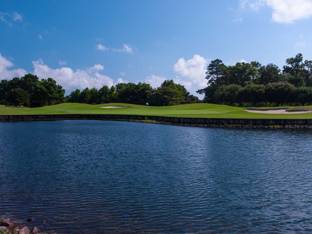 A golf course green with a flag, backed by trees, across a blue lake under a partly cloudy sky.