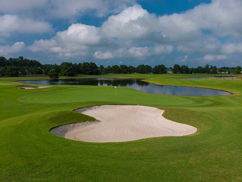 A green golf course with a sand trap, a pond, and a flag on the green under a cloudy sky.